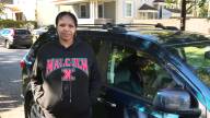 A woman in hoop earrings and a black hoodie that says “Malcolm X” stands in front of her car