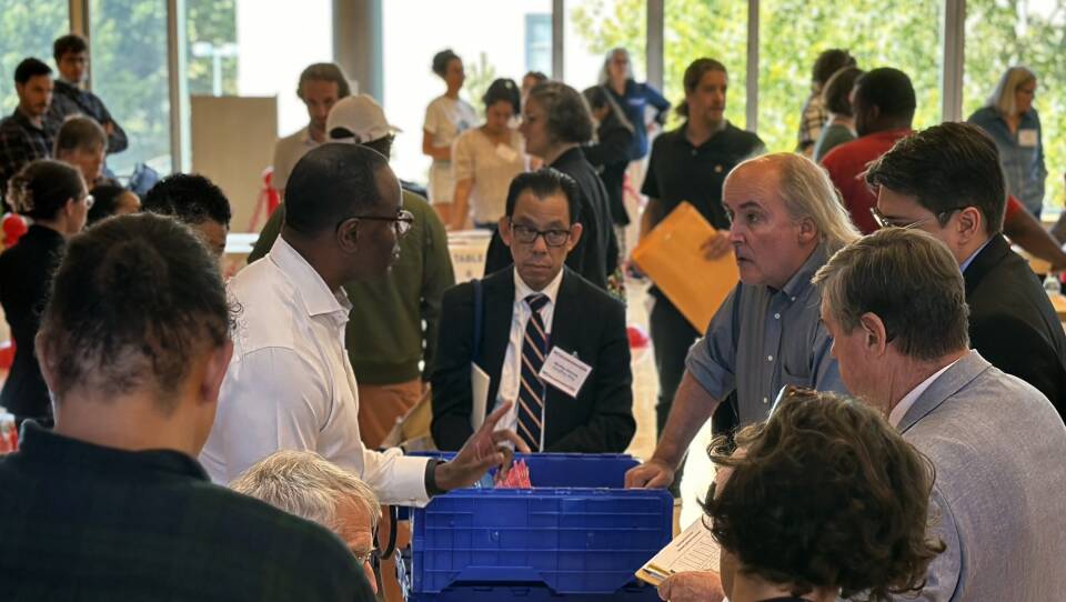 Two people appear to be arguing as they stand by a table with a large plastic box full of papers. A handful of other people are watching. The room is crowded with other people.