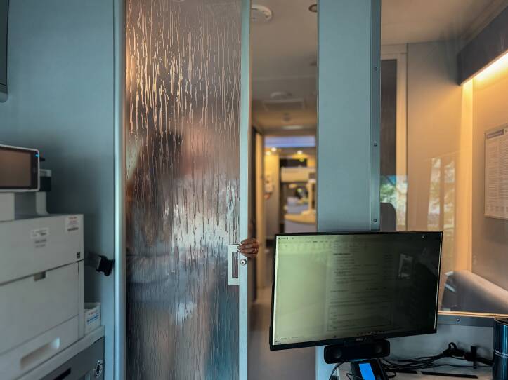 A person is seen in silhouette behind a semi-transparent privacy screen dividing a clerical space from a testing space in a medical clinic.