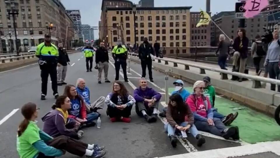 A group of protestors sitting in the road. Behind them is a group of police officers.