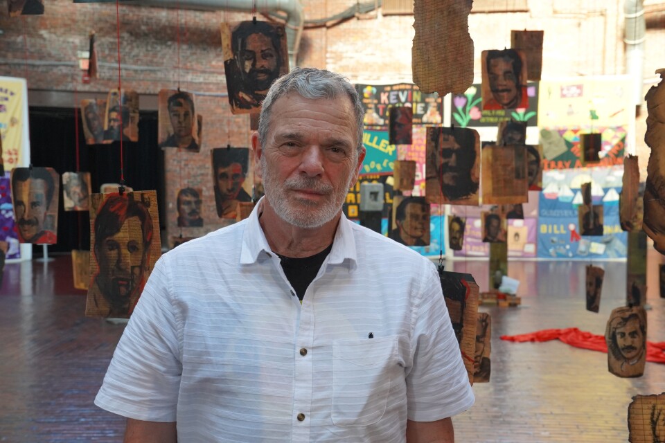A man stands in the middle of a large art exhibition. Dozens of portraits of people who have died from complications from HIV/AIDS hang in the background
