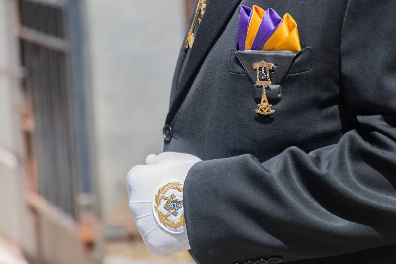 A closeup of a man wearing a white glove with a gold and blue Masonic symbol and the pocket square pin with the Masonic symbol.