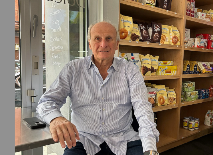 A man in a pinstriped dress shirt poses for a portrait sitting at a counter next to the grocery shelves in a cafe.