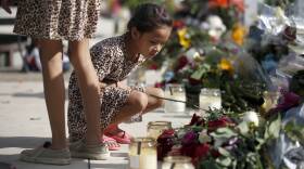 A young child wearing a dress and sandals squats in front of candles and flowers
