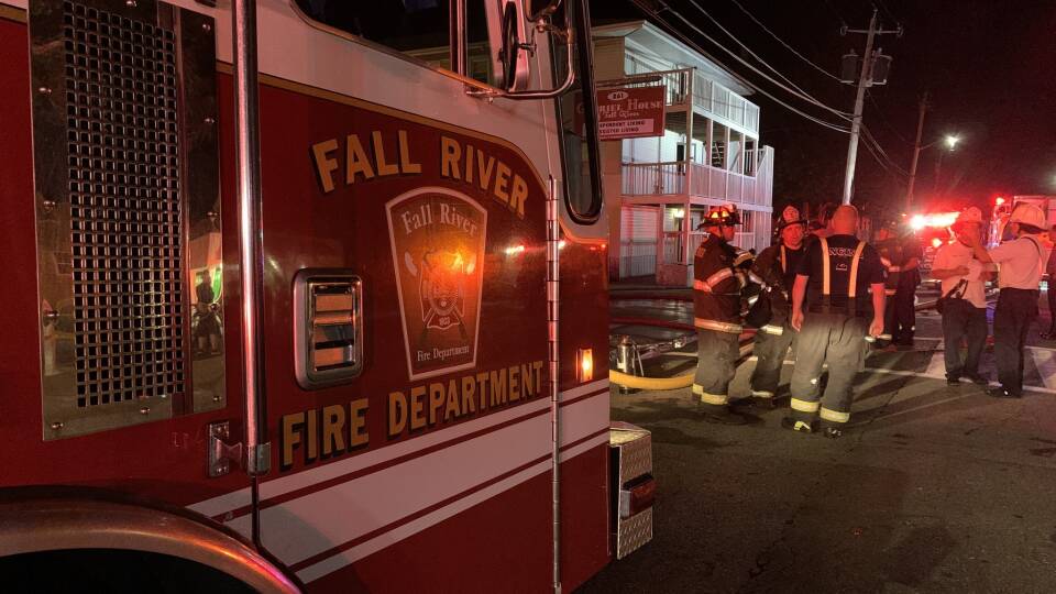 A group of firefighters talk to one another between firetrucks in the street, with their flashing lights glowing.